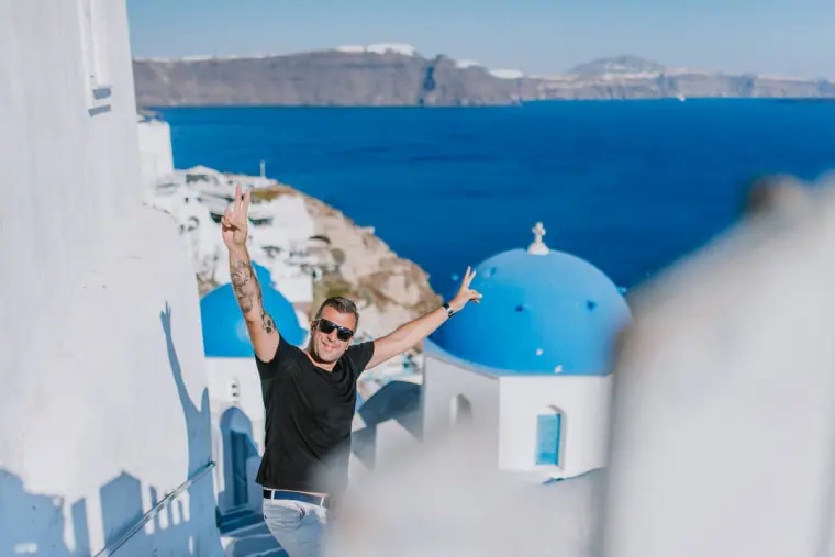 Man in sunglasses with arms raised beside a blue dome above the caldera