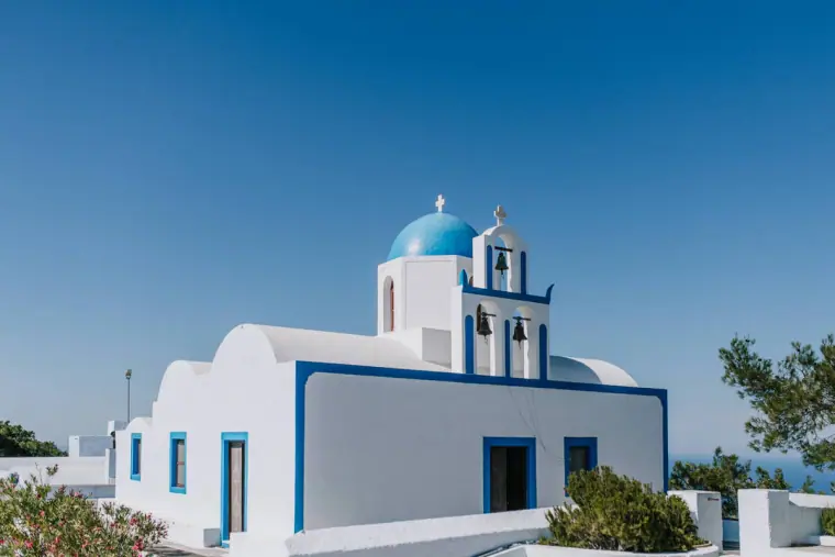 White church with a blue dome and bell tower under a clear blue sky in Santorini