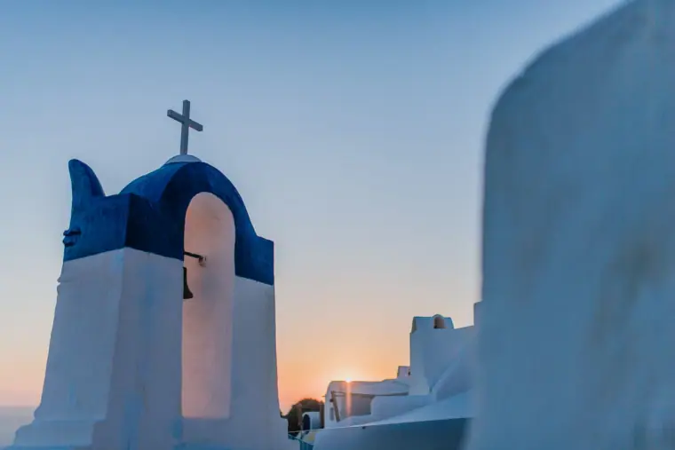 Blue-and-white bell tower with a cross silhouetted at sunset