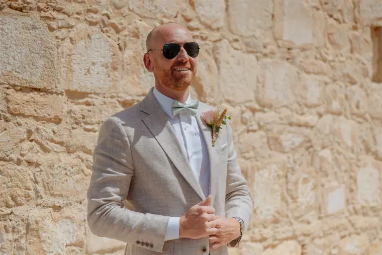 Ben in sunglasses and a beige suit adjusting his jacket against a stone wall