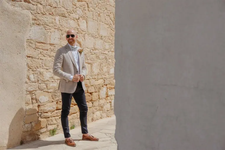Groom in sunglasses standing beside a sunlit stone wall