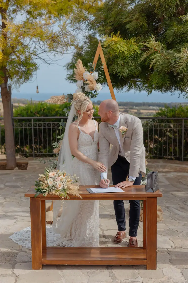 Couple signing the register at a wooden table beneath a triangular boho arch