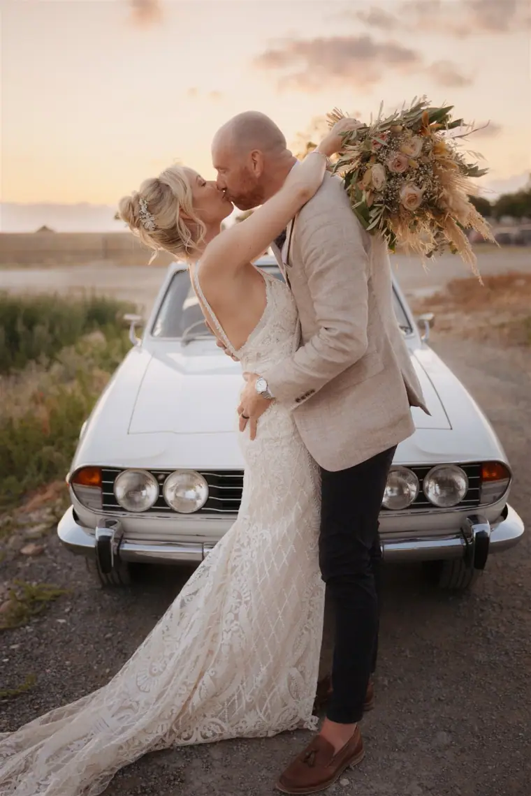 Couple kissing beside the vintage Triumph convertible at sunset
