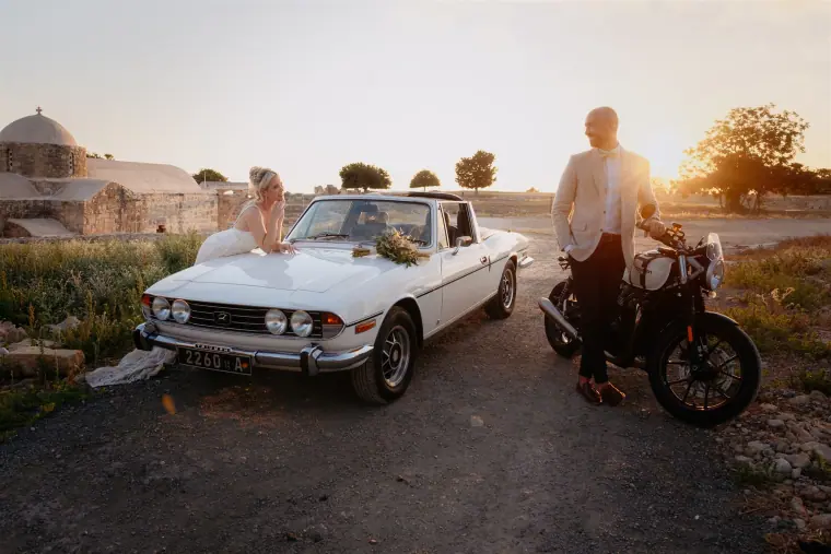 Bride on the bonnet of a white convertible as the groom leans on a motorbike at sunset