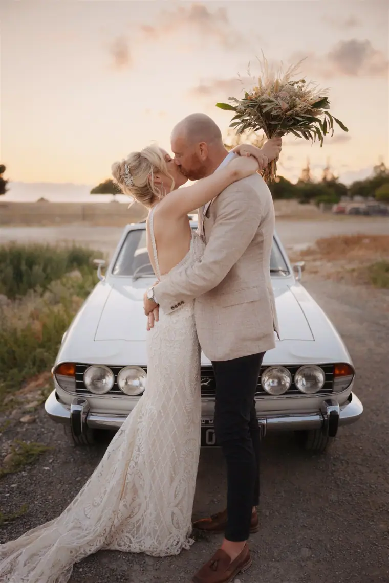 Groom kissing his bride beside the vintage car, her bouquet raised