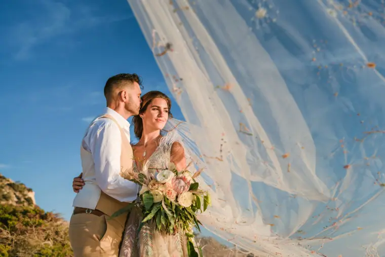 Groom kissing his bride's temple as her veil billows against the blue sky
