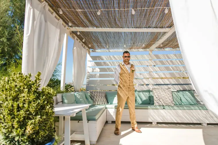 Groom in a gold waistcoat standing under a white-draped reed canopy