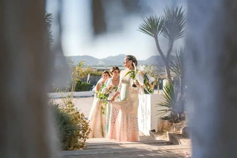 Bride walking out with two companions in pale gowns, seen between drapes