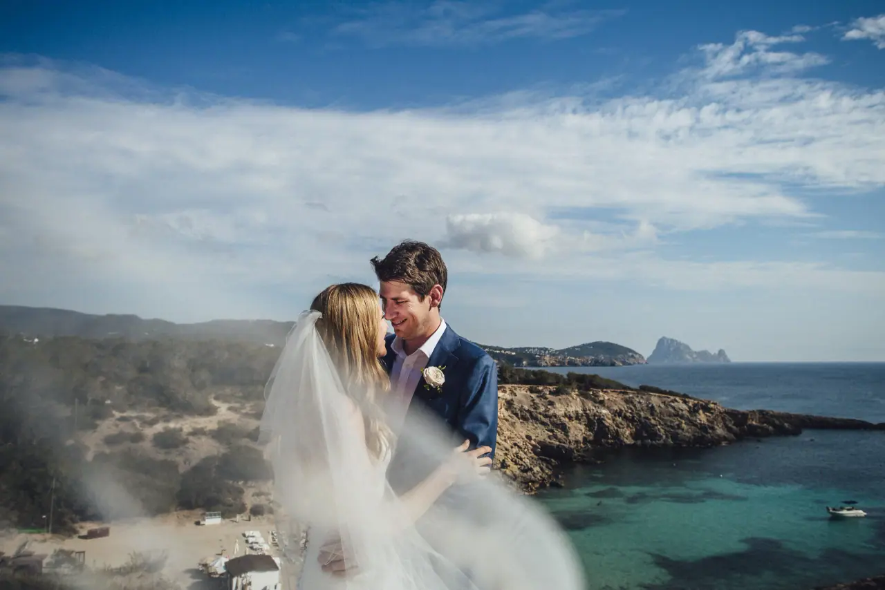 Stephanie and Paul embracing on a clifftop above a turquoise cove in Ibiza