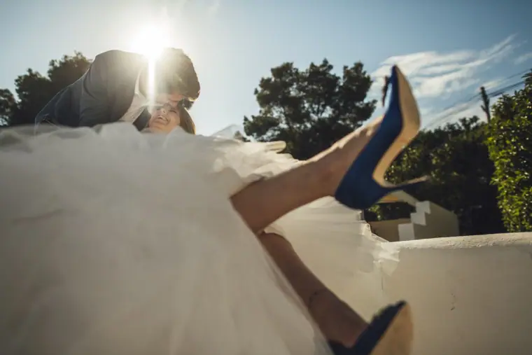 Groom kissing his bride as her blue shoes kick up, sun flaring behind