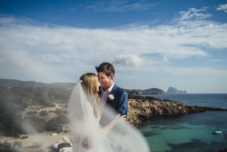 Stephanie and Paul embracing on a clifftop above a turquoise Ibiza bay, veil blowing in the wind