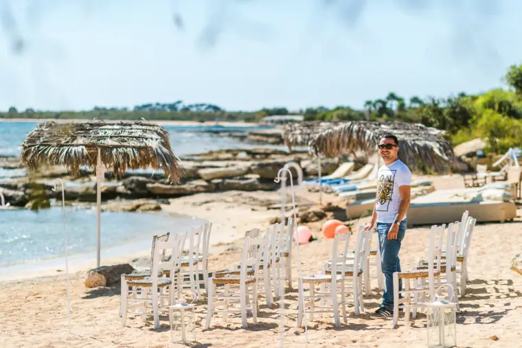 White ceremony chairs set out on the sand beside thatched parasols as a guest looks on