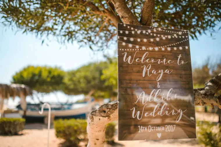 Rustic wooden welcome sign for Raya and Adhib's wedding hanging from a pine tree with fairy lights