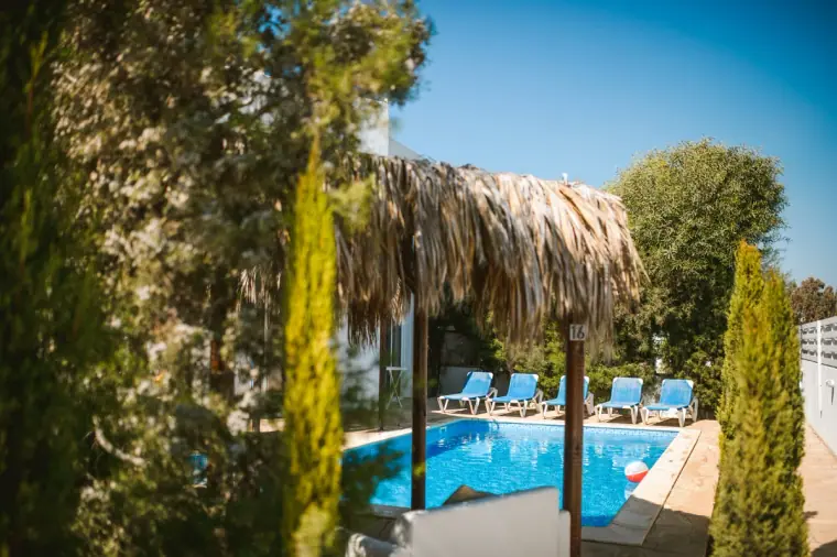 Swimming pool with blue loungers and a thatched parasol among cypress trees at the villas