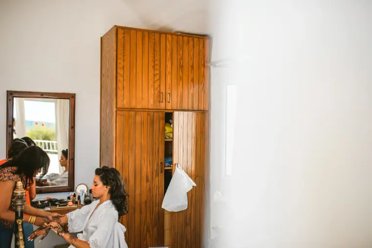 Bride in a white robe having her make-up done by a mirror beside a wooden wardrobe