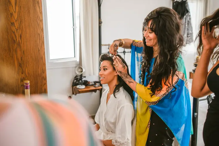 Woman in a bright sari-style wrap fixing the bride's hair as she sits in her robe