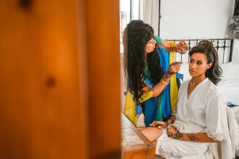 Bride in a white robe sitting on the bed as a woman in a blue and yellow wrap adjusts her hair