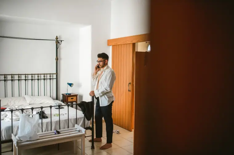 Groom in a white shirt on the phone in a villa bedroom with a wrought-iron bed