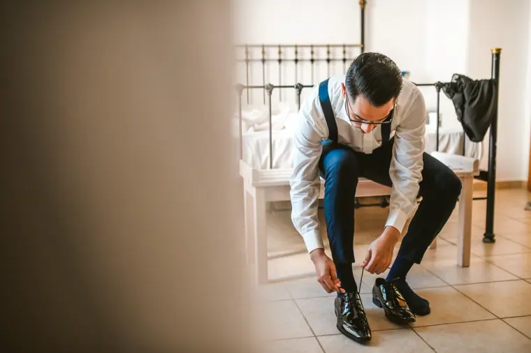 Groom in a white shirt and navy trousers tying his shoes beside the bed