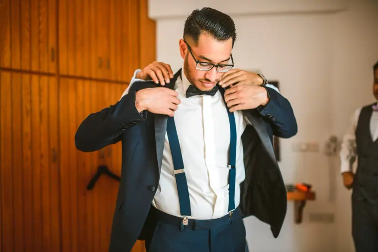 Groom in a navy tuxedo and braces pulling on his jacket in the villa bedroom