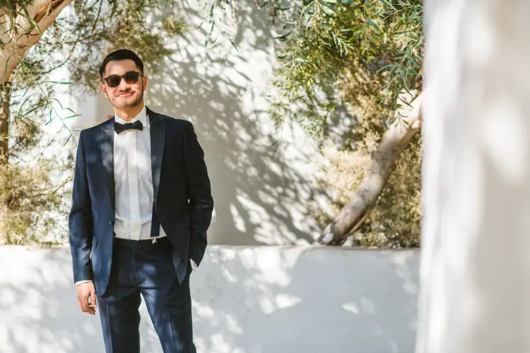 Groom in a navy tuxedo and sunglasses standing by a white wall under olive branches