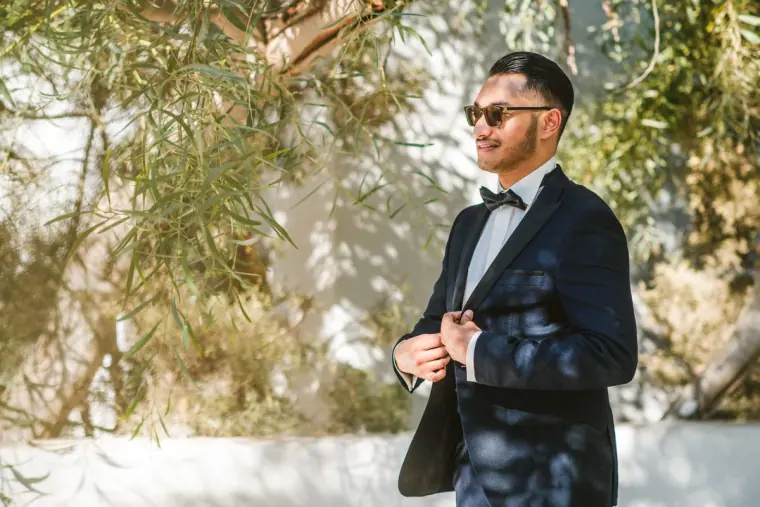 Groom in sunglasses fastening his tuxedo jacket in dappled shade, Thalassines Beach Villas