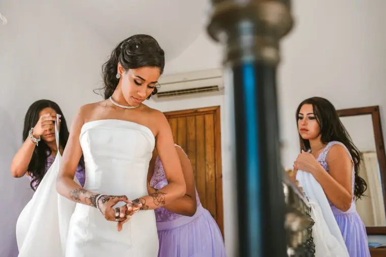 Bride in her strapless gown fastening a bracelet as bridesmaids in lilac get ready
