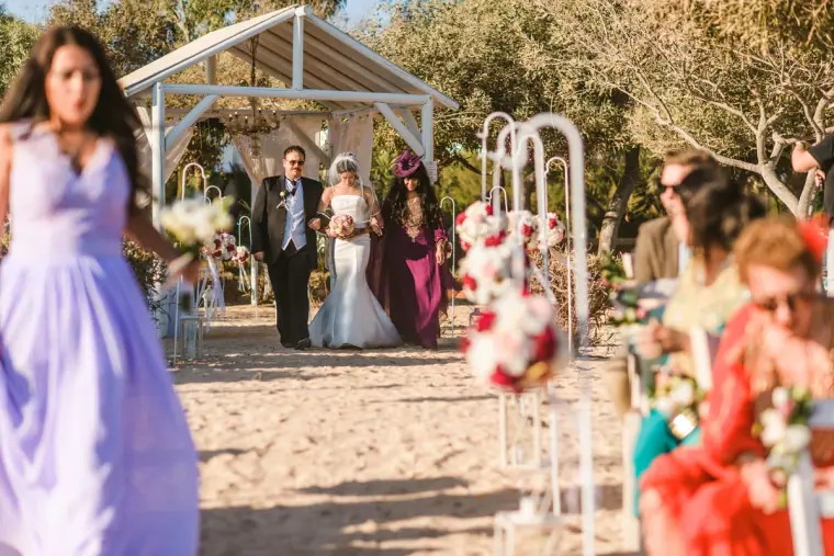 Bride escorted down the sandy aisle towards the white gazebo as guests watch