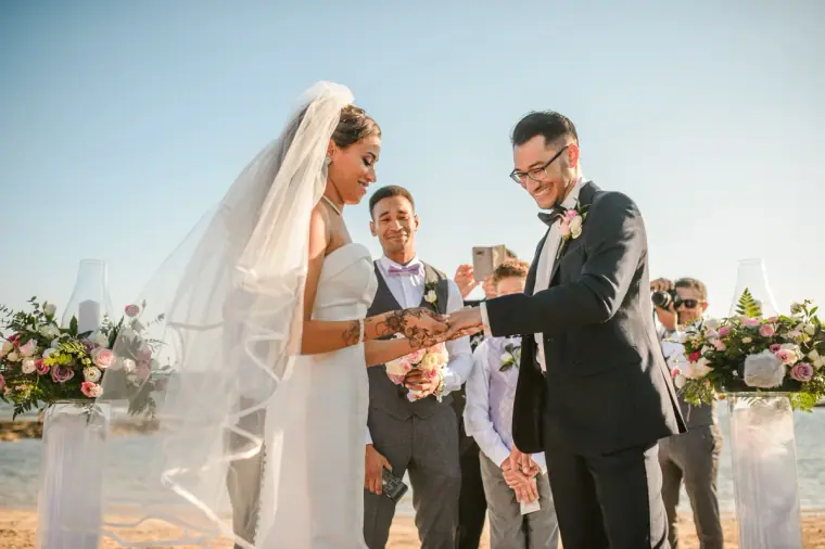 Bride and groom exchanging rings beneath a bright sky by the sea