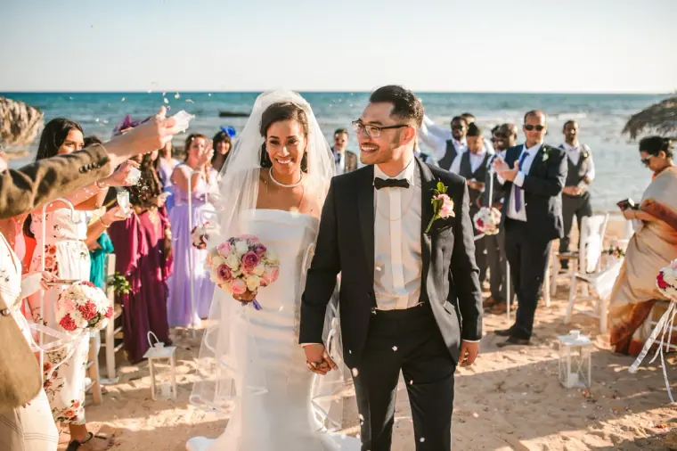 Bride and groom smiling as guests shower them with confetti on the sand
