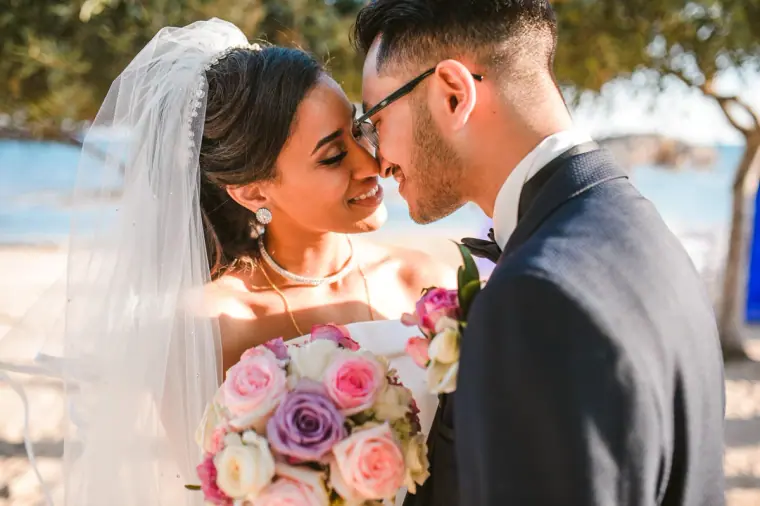 Raya and Adhib touching noses, the bride holding pink and purple roses by the sea