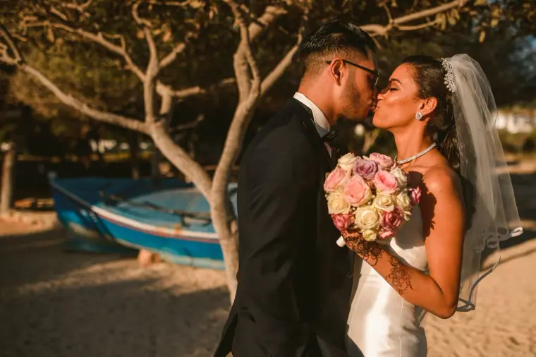 Bride and groom kissing under a pine tree with a wooden boat behind, Thalassines Beach Villas