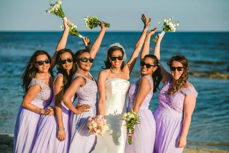 Bride and bridesmaids in lilac raising their bouquets and cheering by the sea