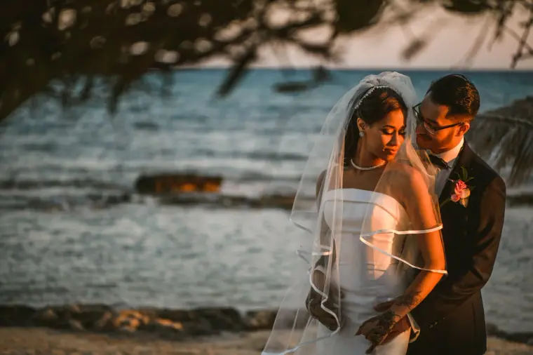 Bride in her veil leaning into the groom beside the sea under a tree