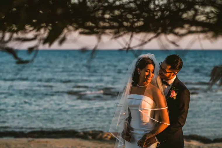 Groom kissing the bride's head as they stand by the water at dusk