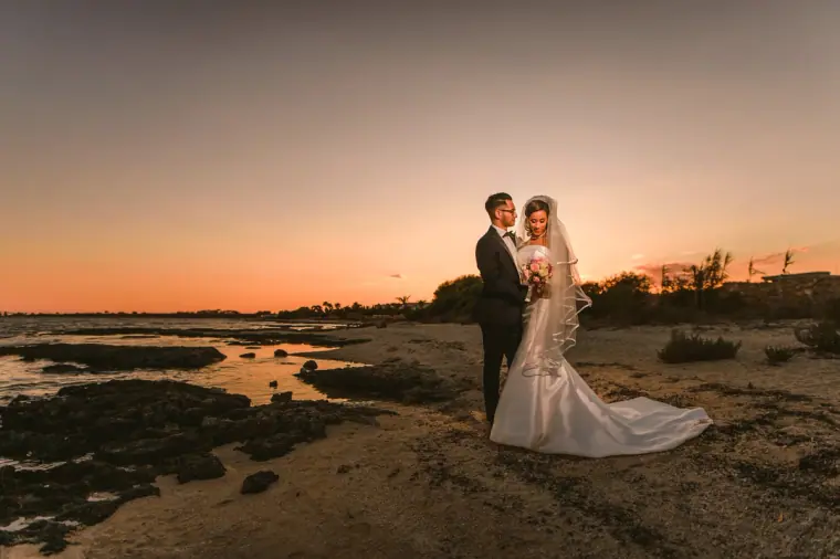 Newlyweds embracing on the beach at sunset, the bride's train spread on the sand