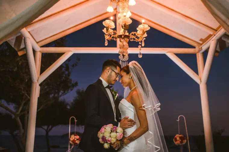 Groom kissing the bride's forehead under the chandelier-lit gazebo at night