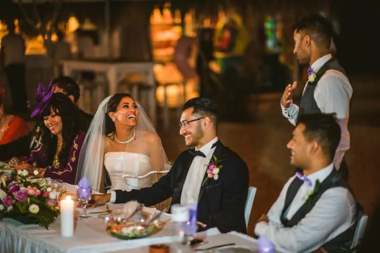 Bride and groom laughing at the top table as a groomsman gives a speech