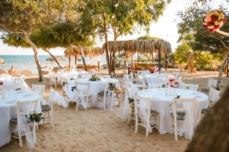 Round tables with white chairs set on the sand beneath pine trees by the sea, Thalassines Beach Villas