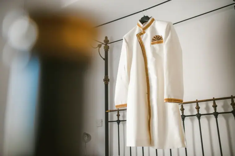 Groom's cream sherwani with gold trim hanging on a wrought-iron bed frame
