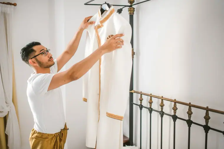Groom in a white T-shirt reaching for his cream sherwani hanging from the bed frame