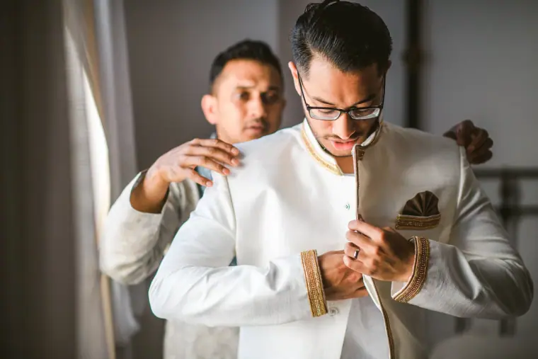 Groom fastening the buttons of his cream sherwani as a relative helps him