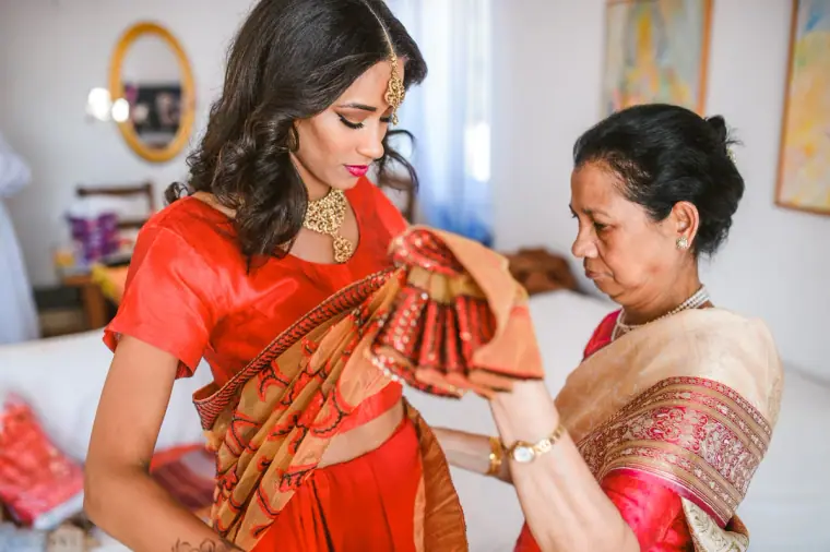 Bride in a red and gold sari having her pallu arranged by an older woman