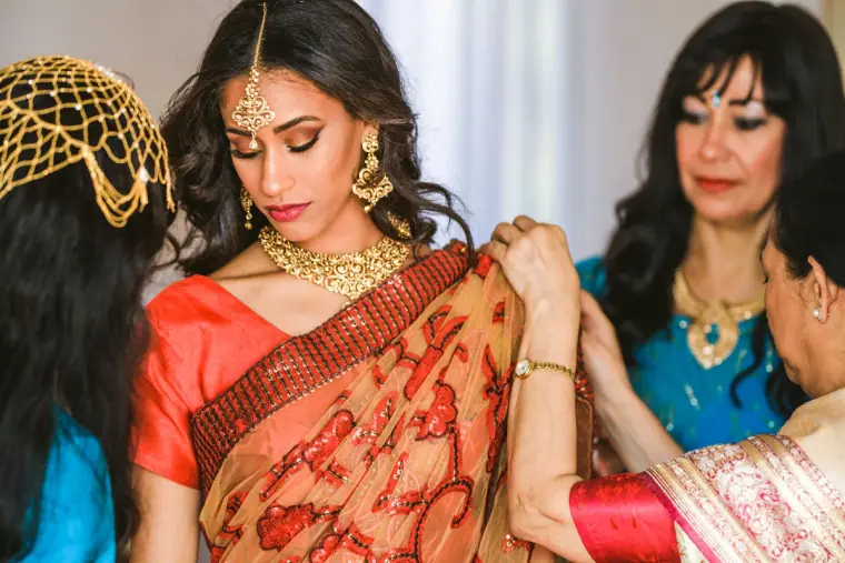 Bride in a gold maang tikka looking down as women adjust her sari