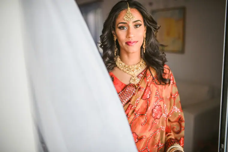 Bride in a red and gold sari and maang tikka framed by a white curtain