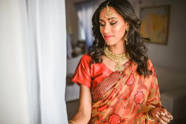 Bride glancing down at her sari by a sunlit curtain in the villa