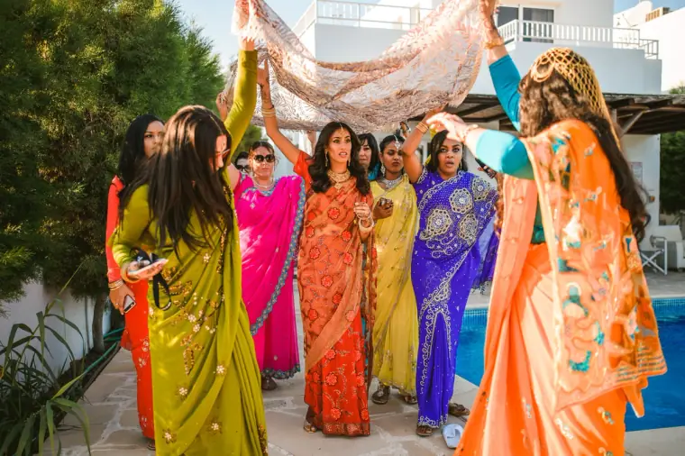 Women in vibrant saris carrying an embroidered canopy over the bride by the pool