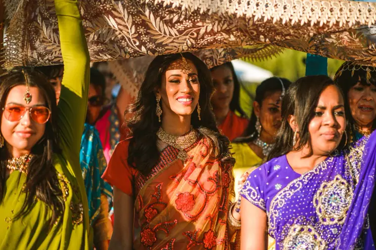 Bride smiling beneath the embroidered canopy, surrounded by women in bright saris