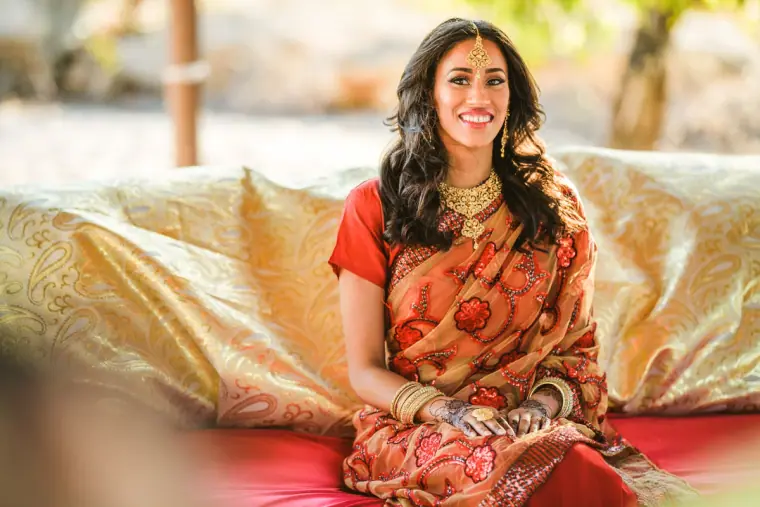 Raya in her red sari seated on gold cushions under the beach canopy