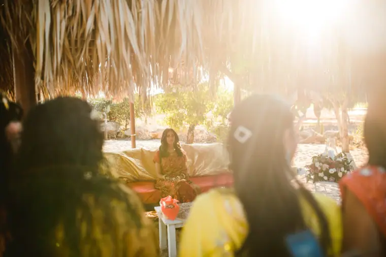 Guests in bright outfits gathered under the thatched canopy as the bride waits on gold cushions